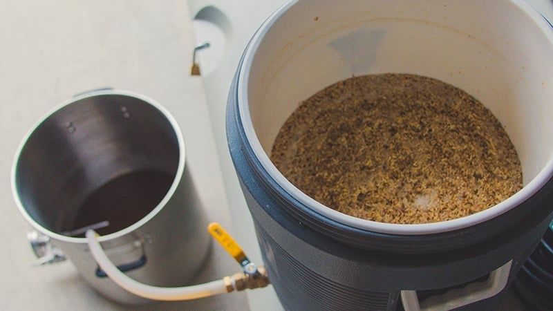 Open homebrew mash tun filled with grain beside a metal bucket